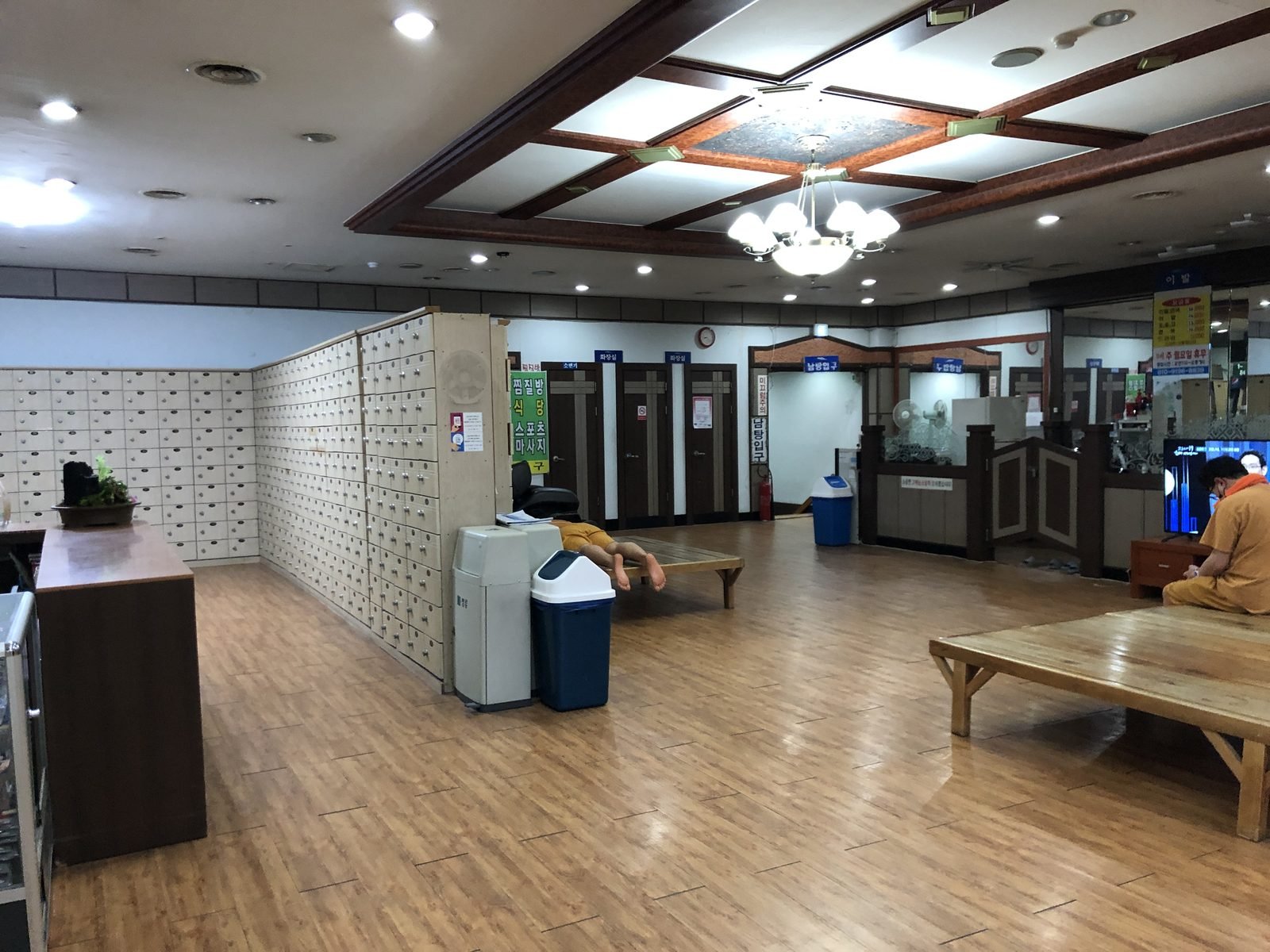 Wide wooden-floored lounge inside a Korean bathhouse complex with wall-to-wall lockers, low platforms and visitors in matching orange house clothes