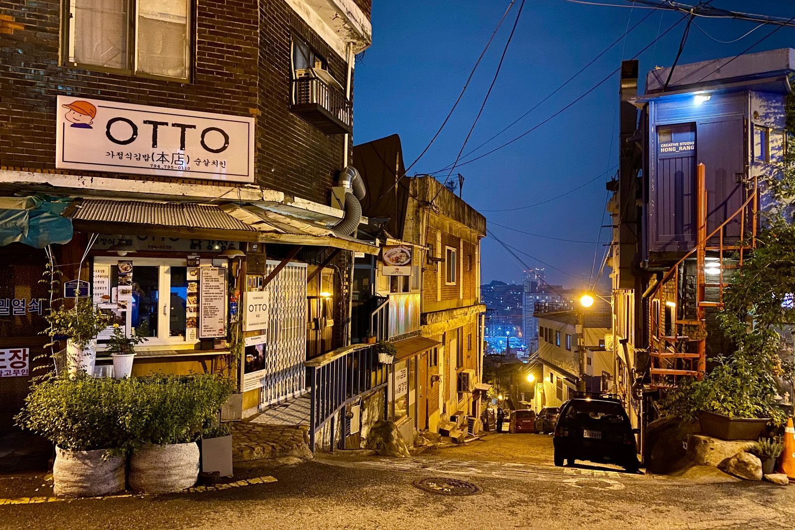 A steep Itaewon side street at dusk with a small shop sign, narrow houses and a parked car