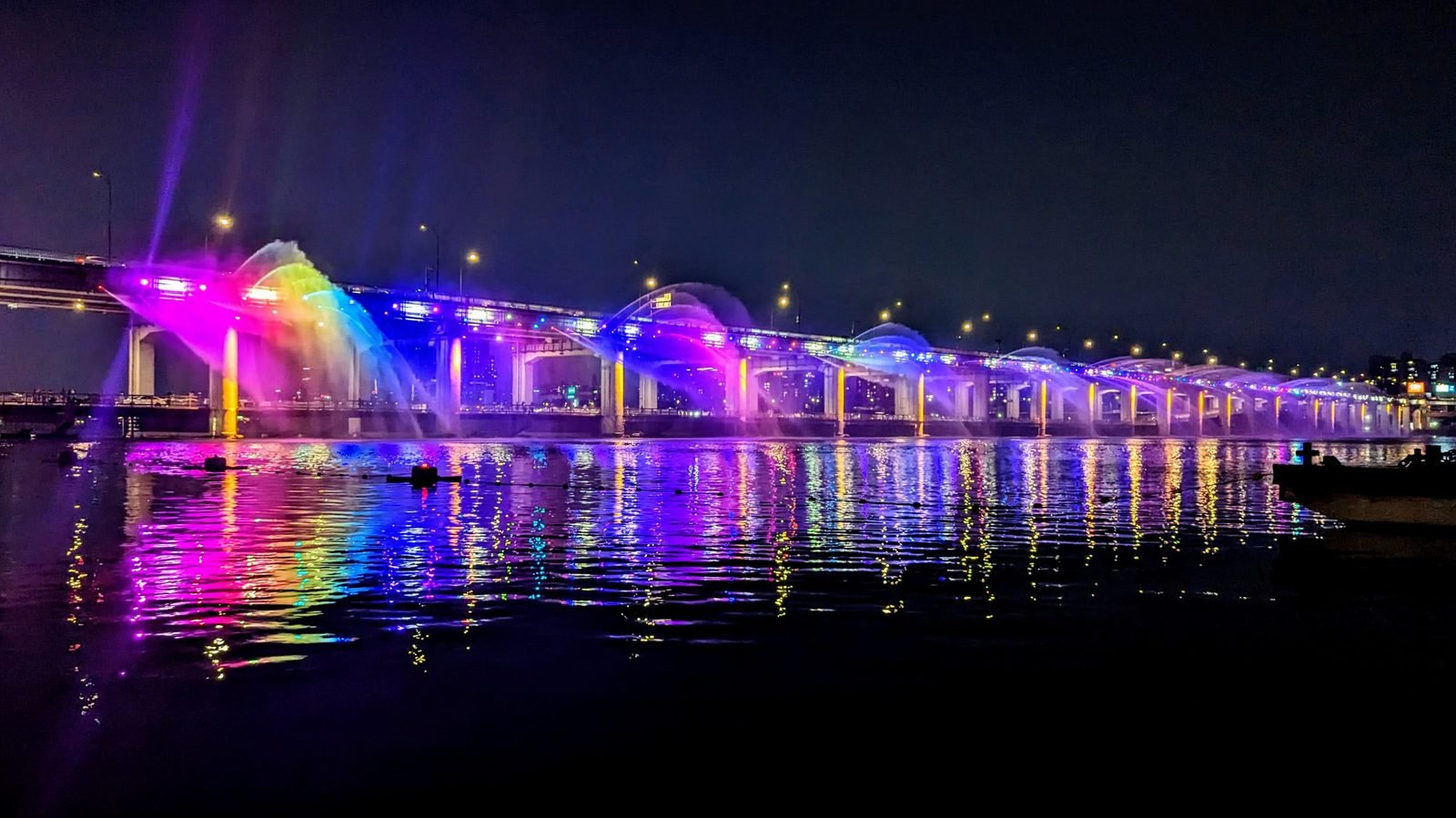 Arcs of water lit in rainbow colors along Banpo Bridge at night, reflected in the Han River